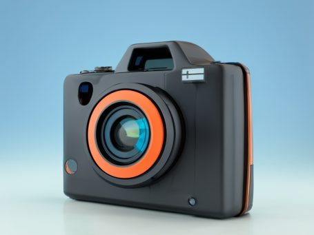 A rendered image of a compact retro camera with a black body and orange accents on the lens and rim.